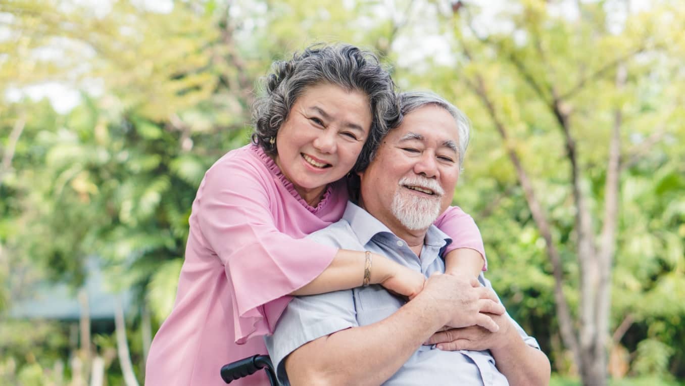 A senior Asian wife happily embraces her smiling husband in his wheelchair in a park