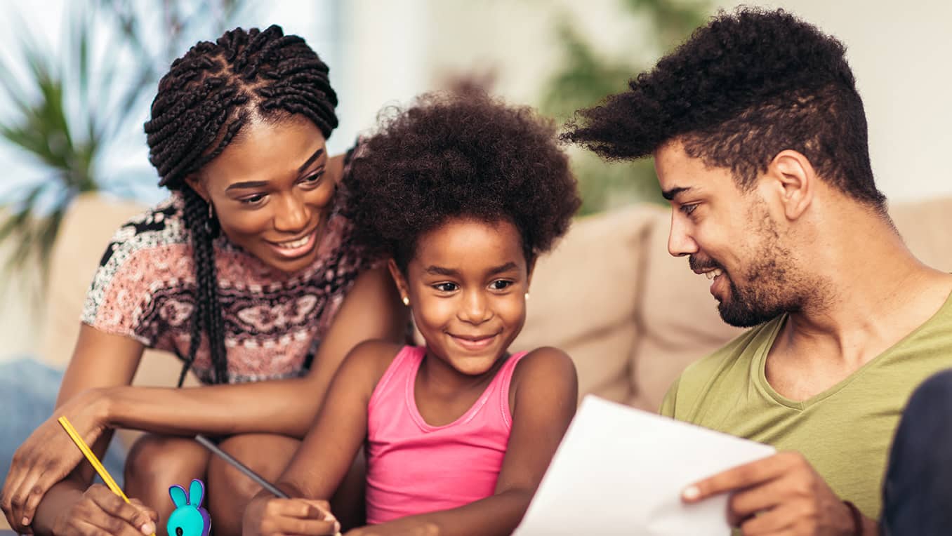 A smiling family of two parents and a child doing a writing activity together at a coffee table
