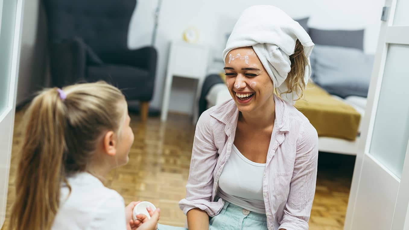 A mother and daughter sit on the floor applying face cream and laughing together
