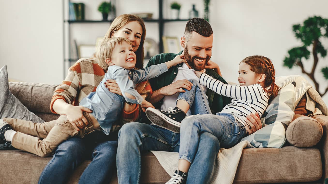 Family plays together on a brown couch in the living room with green plants in the background